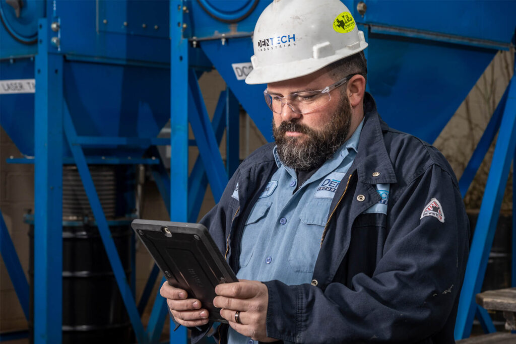 An OSHA dust compliance expert in a hard hat and protective goggles, holding a tablet.