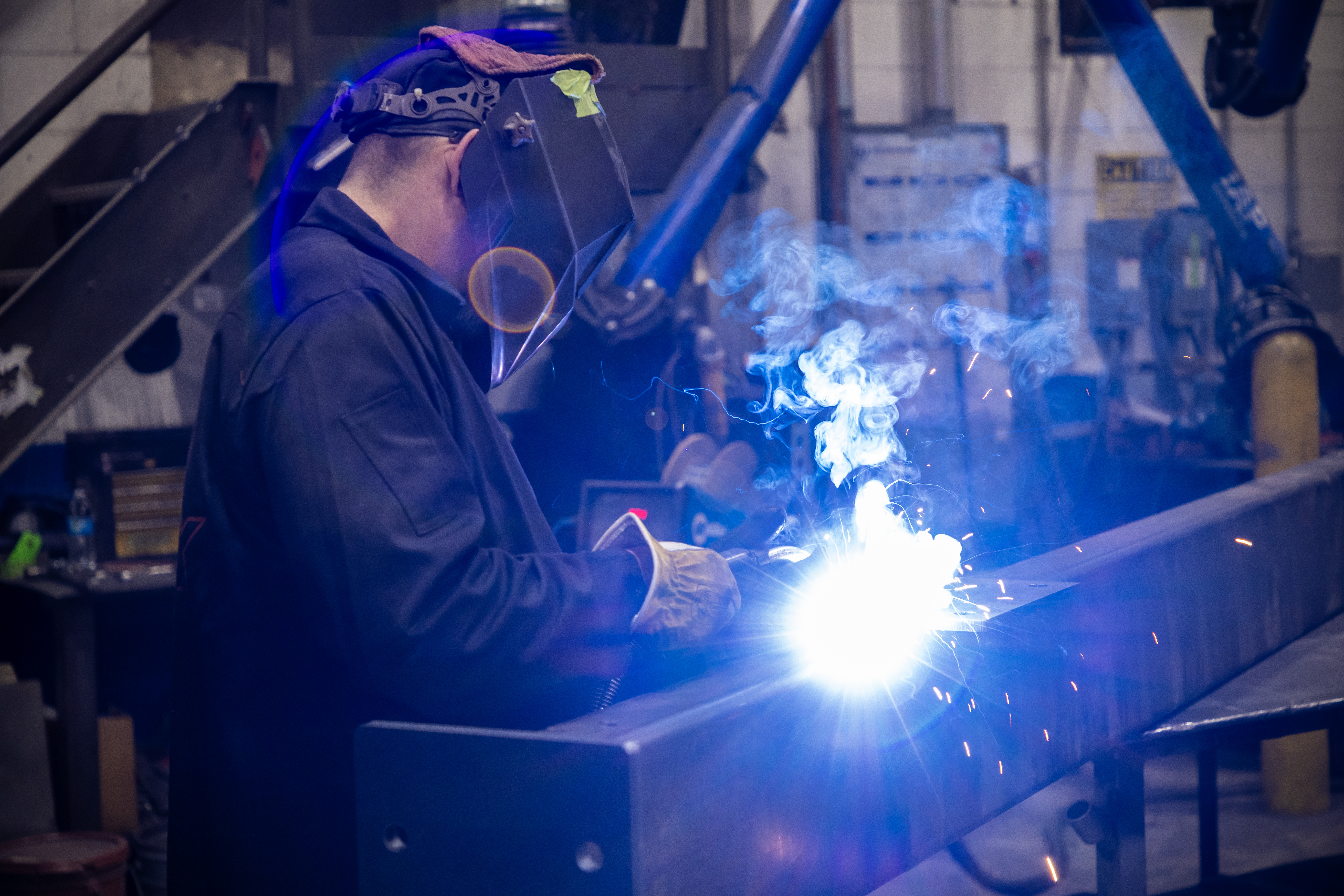 A person wearing a welding helmet is welding on a large metal beam.