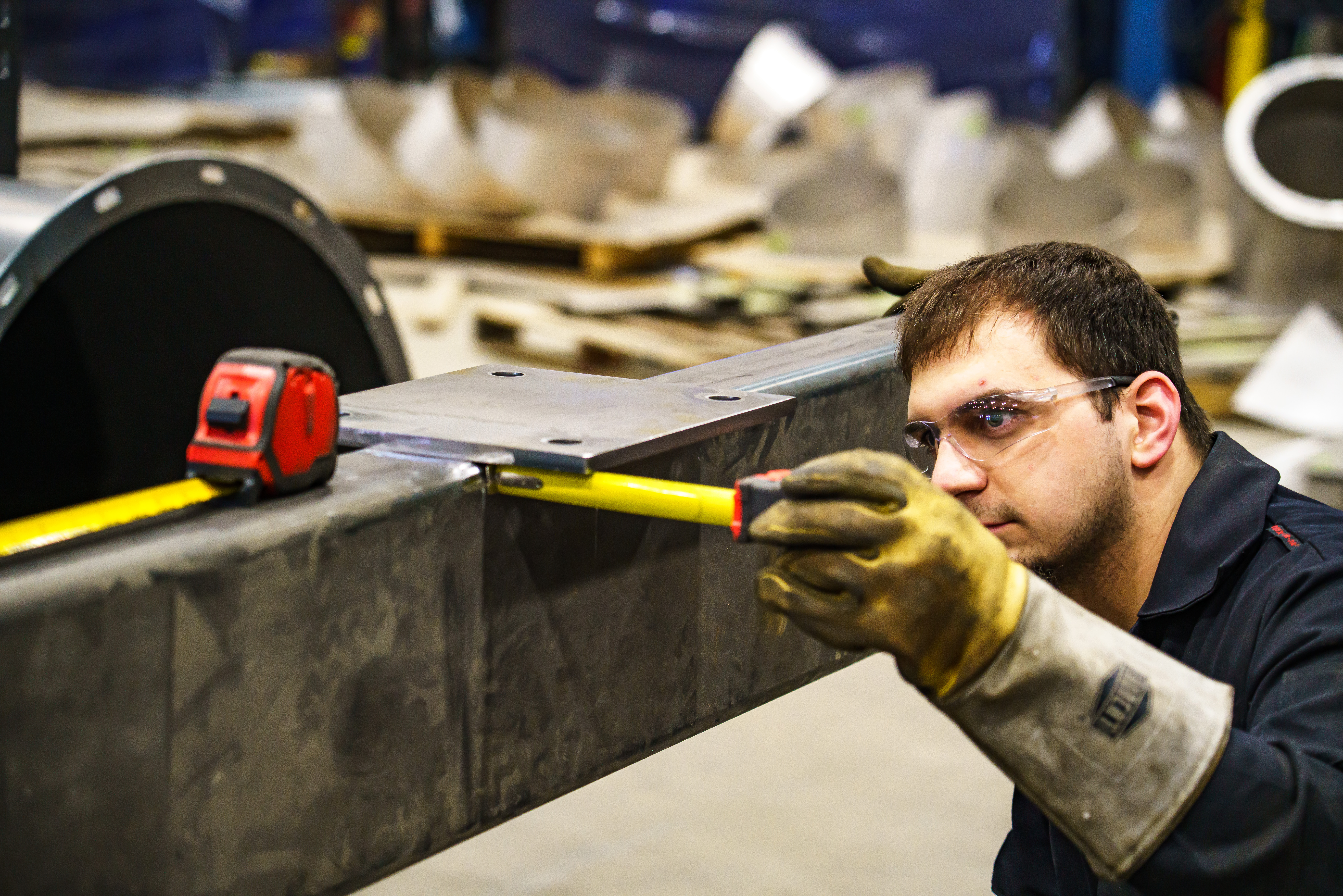 A person wearing safety glasses and protective loves uses a tape measure to measure a piece of flat metal.