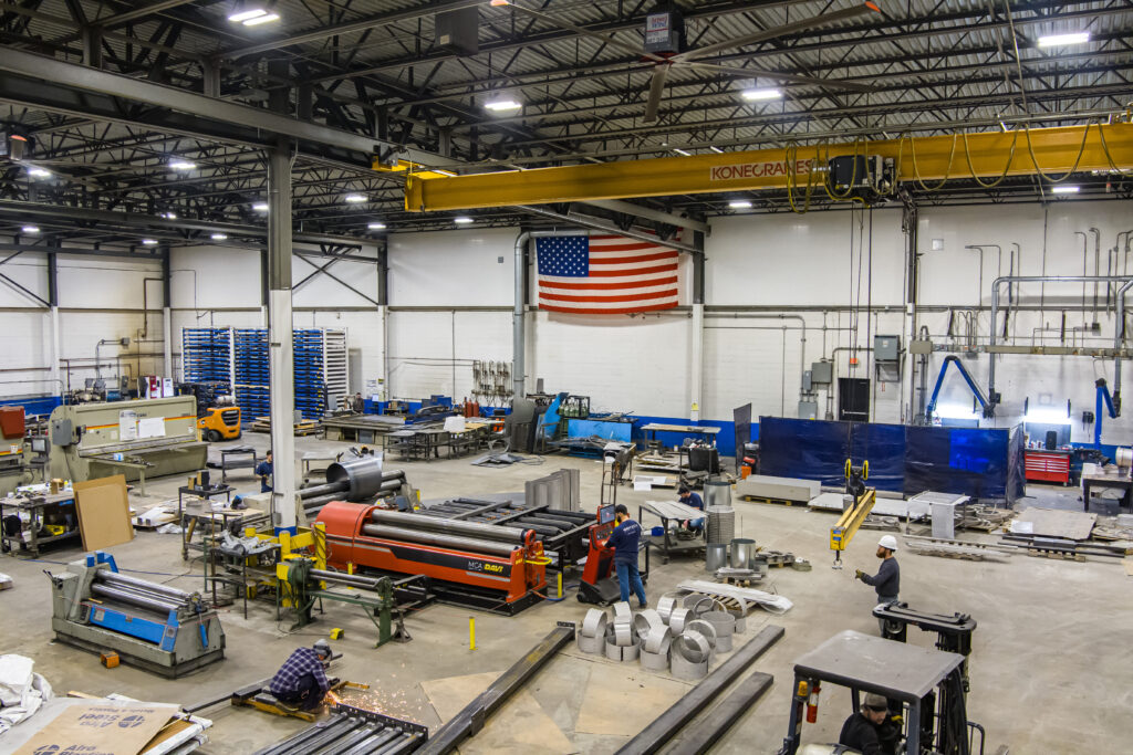 A fabrication shop with various equipment arranged on the floor. Several employees are working with equipment, including an electric grinder, a machine that rolls metal, an overhead crane, and a forklift.