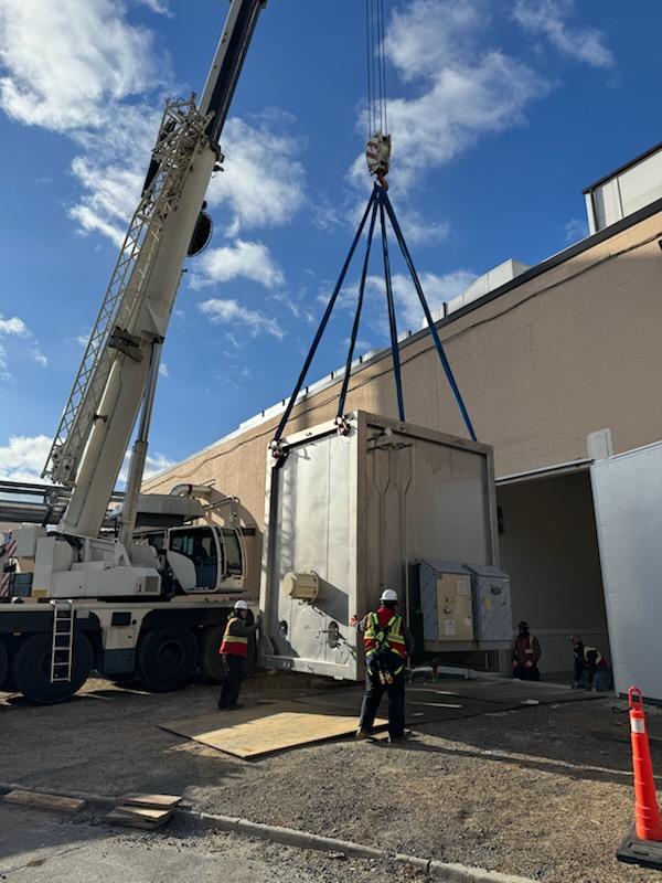 A crane holds a large piece of equipment at the opening to a building. Four people align the equipment to enter the building.