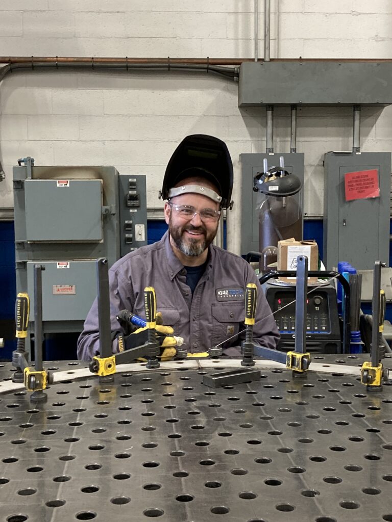 A man sits at a welding station. He's wearing a helmet and safety glasses. A piece of metal is bolted to the table in front of him.