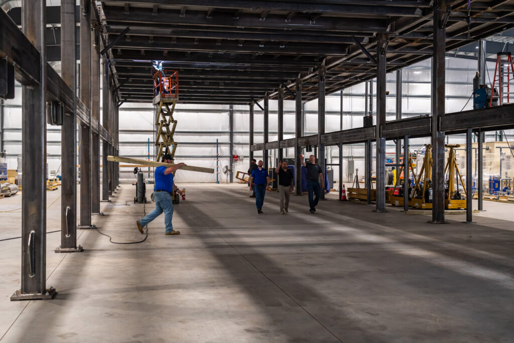 Factory floor with a man carrying a wood beam, three men walking, and a construction lift in the background.