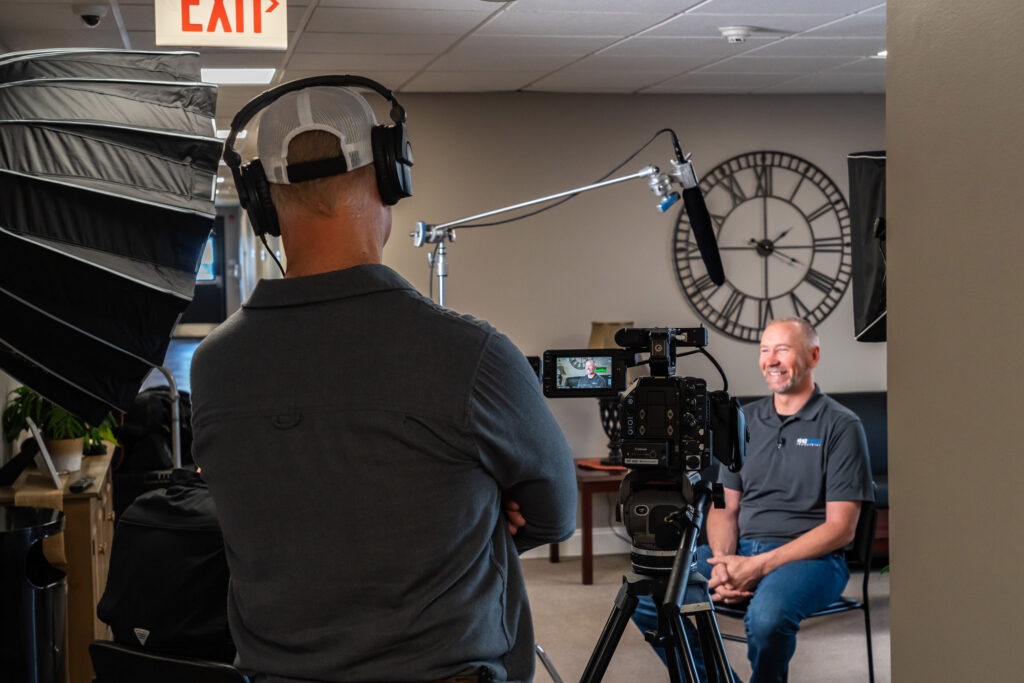 A man sits on a chair in front of a camera and mic for an interview. A second man wearing headphones watches from behind the camera.