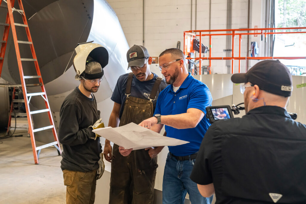 A man with a video camera records three men who are looking at a paper together. Large ductwork and a ladder are in the background.