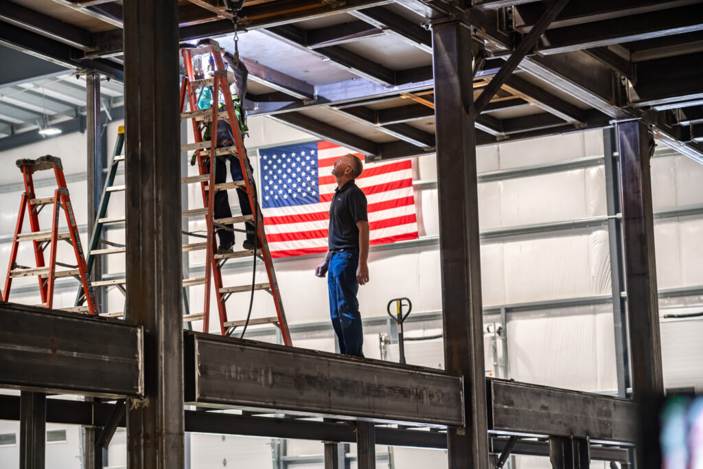 A man is standing on a metal platform and looks up at a welder working at ladder height.