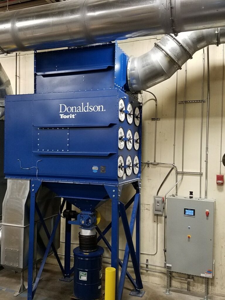 To select the proper dust collector, consider the application and processes in your facility. This dust collector is installed inside a building. The system includes a sealed drum kit below the collector, ductwork, and an electrical control panel.